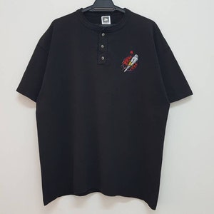 May include: Black short-sleeved t-shirt with a button-up collar and a small embroidered design on the front. The design features a figure in a red circle with the text "The Real Deal" above it.