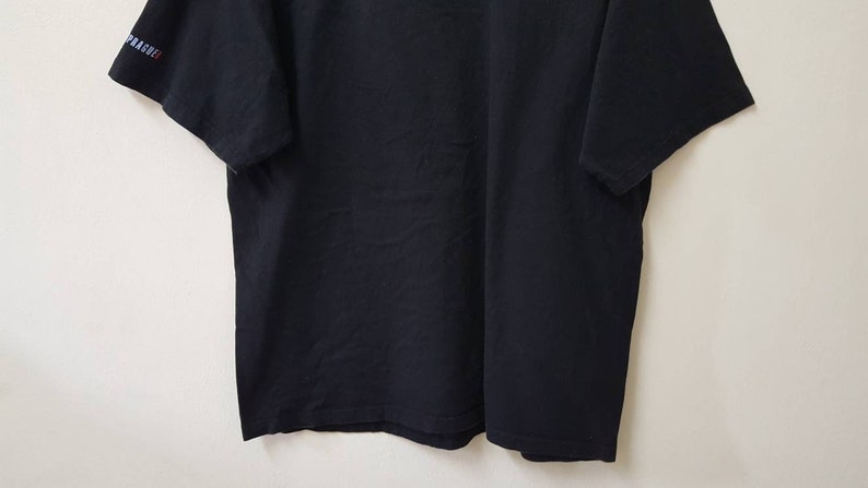 K&ouml;nnte beinhalten: Ein schwarzes T-Shirt mit kurzen &Auml;rmeln. Der &Auml;rmel tr&auml;gt das Wort "PRAGUE" in blauen und roten Buchstaben. Das T-Shirt ist aus einem weichen, dunklen Stoff gefertigt und wird vor einem schlichten wei&szlig;en Hintergrund pr&auml;sentiert.