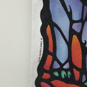 Puede incluir: Una camiseta blanca con un dise&ntilde;o de mariposa de vitral de colores. El texto "&copy; 1993 LIQUID BLUE" est&aacute; impreso en la esquina inferior derecha de la camiseta.