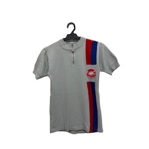 May include: A light gray short-sleeve cycling jersey with a zipper at the collar. The jersey features red and blue vertical stripes on the right side and a circular logo patch. The jersey is hanging on a black hanger.
