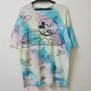 Może przedstawiać: Koszulka tie-dye z kreskówkowym wizerunkiem Myszki Miki prowadzącej statek kosmiczny. Koszulka ma jasnoniebieski, różowy i żółty schemat kolorystyczny.