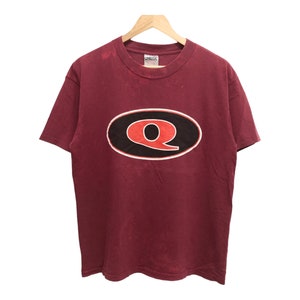 May include: A maroon t-shirt featuring a large black oval with a red "Q" in the centre. The shirt is made from a soft, comfortable cotton blend.