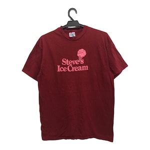 Op de afbeelding: Een roodbruin T-shirt met korte mouwen met een roze ijshoorn-afbeelding en de tekst "Steve's Ice Cream".