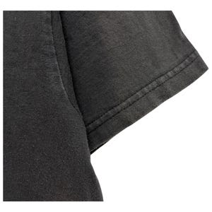 Puede incluir: Primer plano de una manga de camiseta gris oscuro. La tela parece ser una suave mezcla de algod&oacute;n, con textura visible y sutiles variaciones de color. El borde de la manga est&aacute; cuidadosamente dobladillado con una costura de doble puntada.