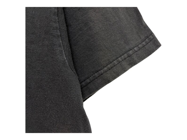 Puede incluir: Primer plano de una manga de camiseta gris oscuro. La tela parece ser una suave mezcla de algod&oacute;n, con textura visible y sutiles variaciones de color. El borde de la manga est&aacute; cuidadosamente dobladillado con una costura de doble puntada.