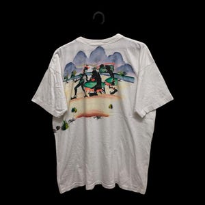 May include: White t-shirt with a colorful graphic on the back featuring figures in a landscape with mountains and a sunset. The shirt is hanging on a black hanger against a black background. The graphic has a vintage style.