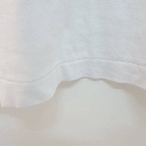 May include: Close-up of the bottom edge of a white t-shirt. The edge is sewn with a straight stitch.