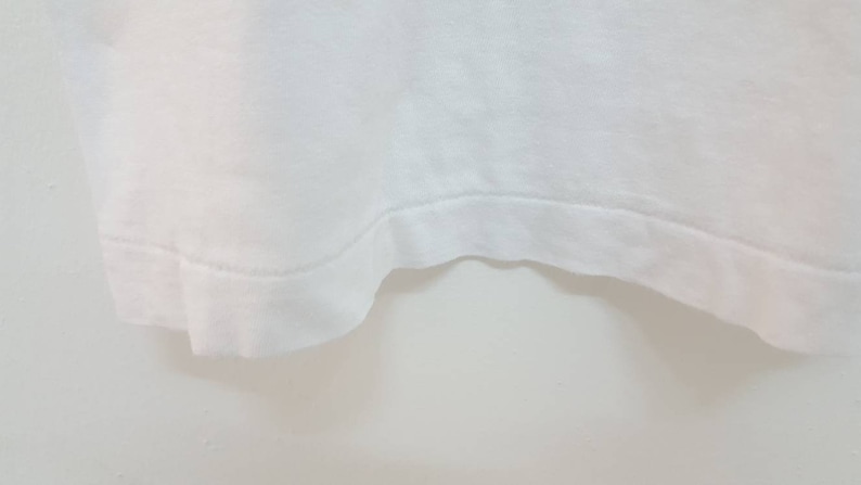 May include: Close-up of the bottom edge of a white t-shirt. The edge is sewn with a straight stitch.