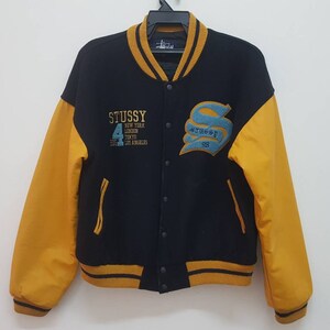 May include: A black and yellow varsity jacket with the word "STUSSY" embroidered on the front. The sleeves are yellow and the body is black with a yellow stripe around the collar and bottom hem.