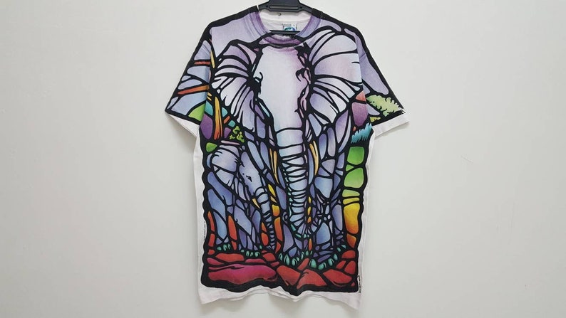 Puede incluir: Camiseta blanca con un dise&ntilde;o de elefantes en vitral de colores. Los elefanten son morados, azules, verdes, amarillos y naranjas.