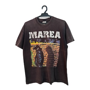 May include: Brown t-shirt with the word "MAREA" in white block letters. The shirt features a graphic of a person in a leather jacket standing in front of a brick wall. The shirt is hanging on a black hanger.