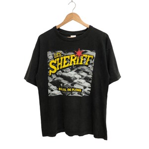 以下が含まれることがあります： 車のシーンと「LES SHERIFF SOLEIL DE PLOMB」というテキストがプリントされた黒いTシャツです。