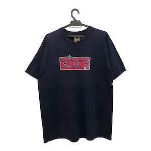 May include: Navy blue t-shirt with a red and white graphic that reads "pimp". The shirt is hanging on a black hanger. The t-shirt is a short-sleeved crew neck.