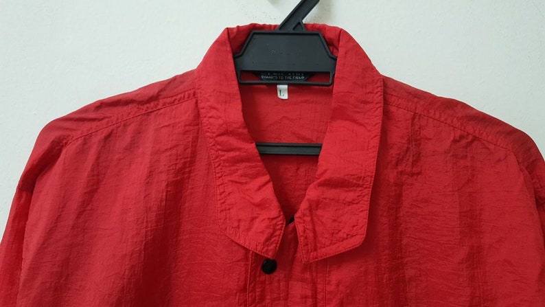 May include: A red long-sleeved shirt with a button-up collar and a single button at the top. The shirt is made of a lightweight, textured fabric.