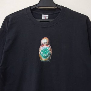 K&ouml;nnte beinhalten: Schwarzes T-Shirt mit einem farbenfrohen Matrjoschka-Puppen-Design. Die Puppe hat ein wei&szlig;es Grinsen und ein Blumenmuster. Das T-Shirt h&auml;ngt an einem schwarzen Kleiderb&uuml;gel vor wei&szlig;em Hintergrund.