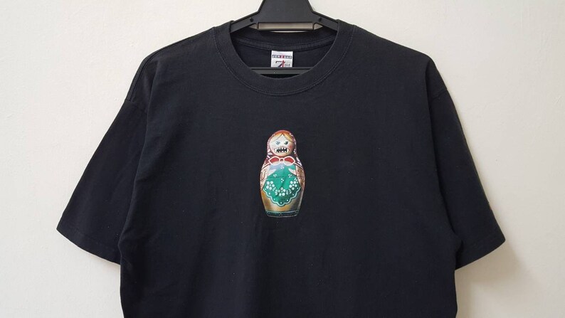 K&ouml;nnte beinhalten: Schwarzes T-Shirt mit einem farbenfrohen Matrjoschka-Puppen-Design. Die Puppe hat ein wei&szlig;es Grinsen und ein Blumenmuster. Das T-Shirt h&auml;ngt an einem schwarzen Kleiderb&uuml;gel vor wei&szlig;em Hintergrund.