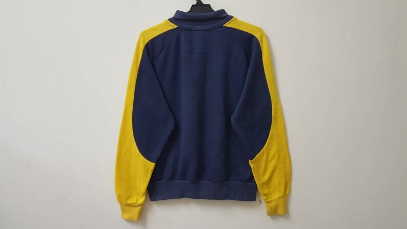 May include: A navy blue sweatshirt with yellow color-blocking on the sleeves. The sweatshirt has a zipper closure and a ribbed collar.