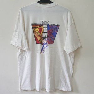 May include: White t-shirt with a graphic of a person riding a bicycle. The graphic is in a triangle shape with the words "Choose Your Weapon" in the center. The words "Team Hits" and "Team L.T.D." are on either side of the triangle.