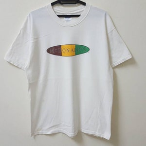 Peut inclure: T-shirt blanc avec un logo ovale brun, jaune et vert qui dit "McDonald's".