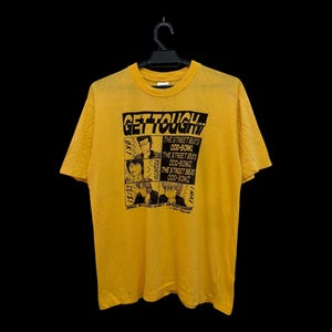 May include: A bright yellow short-sleeve t-shirt featuring a black graphic print on the chest. The design includes the text GET TOUGH, four panels with illustrations of characters, and the repeated text THE STREET BEATS ODD-BOWZ.