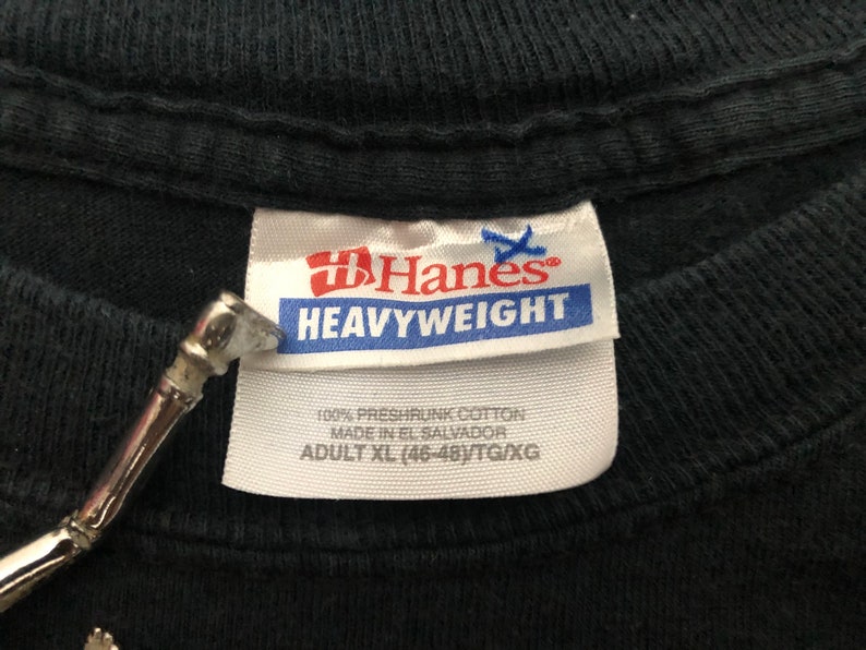May include: A black Hanes Heavyweight t-shirt tag with white text. The tag says "100% preshrunk cotton, made in El Salvador, Adult XL (46-48)/TG/XG".
