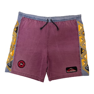 May include: Maroon shorts with an elastic waistband and black drawstring. The sides feature a yellow and black geometric pattern. A circular patch with a red star and a rectangular patch with a red and white logo are on the front panels.