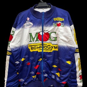May include: A blue and white zip-up athletic jacket featuring the text MOG with a red apple graphic, Technogym branding in yellow, and Nalini Fausto Coppi logos. The jacket has a blue zipper, ribbed collar, and scattered red and yellow diamond shapes.