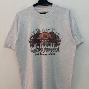 May include: A light gray t-shirt featuring a graphic design with the word "Jaballa" in a stylized font. The design includes a skull, a ship, and the text "Ride the Adventure". The shirt is hanging on a black hanger.