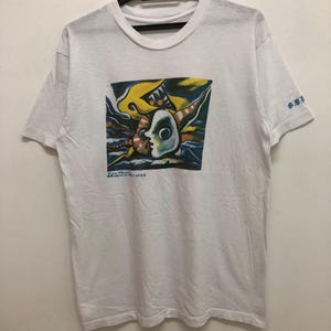 May include: White t-shirt with a colorful abstract graphic print of a face and a yellow, blue, and orange design. The text "Tom Clancy's  Museum of  Art  OPEN" is printed below the graphic.