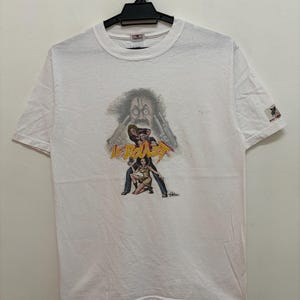 May include: White t-shirt featuring a graphic of a man with guns and two figures. The text "Le Baiser" is printed in yellow and red. The shirt is hanging on a black hanger.