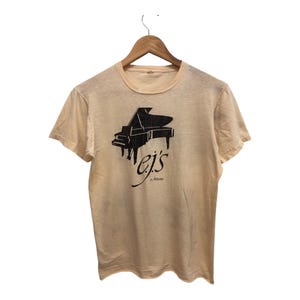 Op de afbeelding: Een lichtbeige t-shirt met een zwarte afbeelding van een vleugelpiano en de tekst "ej's" in cursief.