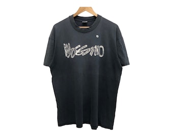 Puede incluir: Camiseta negra con la palabra "Mossimo" en fuente blanca desgastada en el pecho. La camiseta está colgada en una percha de madera. La tela parece desgastada. El fondo es blanco.