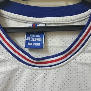 May include: White mesh basketball jersey with blue and red trim around the neck. The jersey has a Champion logo on the front and back. The tag reads "Authentic Athletic Apparel Made in Korea."