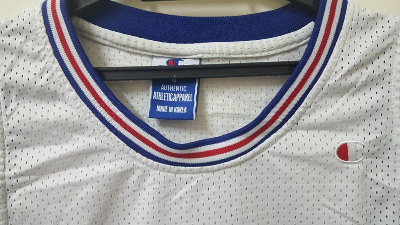 May include: White mesh basketball jersey with blue and red trim around the neck. The jersey has a Champion logo on the front and back. The tag reads "Authentic Athletic Apparel Made in Korea."