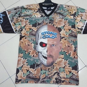 Puede incluir: Una camiseta de estampado de camuflaje con una imagen dividida de una calavera y la cara de Stone Cold Steve Austin. El texto "Stone Cold" está impreso en azul en la calavera. Las mangas tienen rayas blancas y negras con el texto "3:16" y "Austin" impreso en ellas.