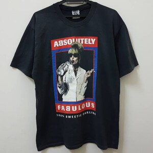 May include: Black t-shirt featuring a graphic of a woman with blonde hair and sunglasses holding a bottle and cigarette. The graphic is in a red, blue and white frame with the text "ABSOLUTELY FABULOUS 100% SWEETIE DARLING".