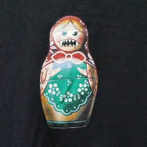 K&ouml;nnte beinhalten: Handbemalte Matrjoschka-Puppe mit grimmigem Gesichtsausdruck. Die Puppe hat ein rotes Kopftuch, eine gr&uuml;ne Sch&uuml;rze mit wei&szlig;en Blumendetails und ein bedrohliches Grinsen. Die Puppe ist vor einem schwarzen Hintergrund platziert.