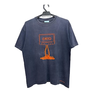 Puede incluir: Camiseta azul oscuro con un gráfico de cinta de cassette naranja. El diseño de la cinta de cassette tiene líneas naranjas que se extienden hacia abajo hasta una pila de líneas naranjas. La camiseta está colgada en una percha negra.