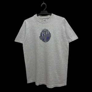 Puede incluir: Una camiseta gris jaspeado con cuello redondo y mangas cortas. La camiseta presenta un gráfico circular en morado y azul con el texto "RB" y "Royal Roots Co." La camiseta está colgada de una percha negra.