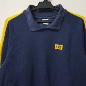 May include: A navy blue long-sleeved polo shirt with yellow stripes on the sleeves. The shirt has a small yellow patch on the chest with the word "NIKE" in white letters.