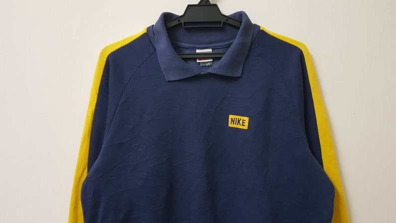 May include: A navy blue long-sleeved polo shirt with yellow stripes on the sleeves. The shirt has a small yellow patch on the chest with the word "NIKE" in white letters.