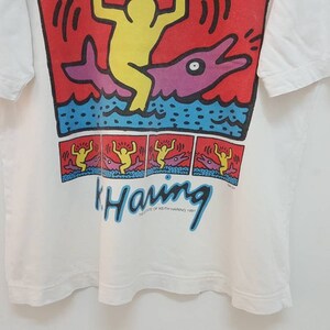 K&ouml;nnte beinhalten: Wei&szlig;es T-Shirt mit einem bunten Grafikdruck einer Person, die auf einem Delfin reitet. Die Grafik enth&auml;lt den Text "Keith Haring" und das Jahr 1984.