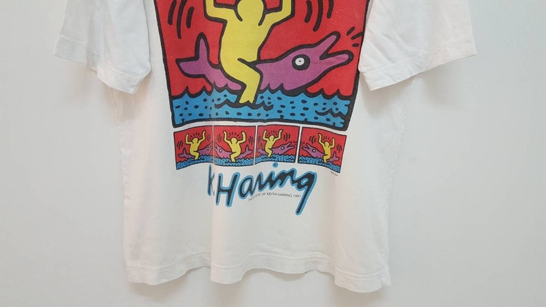 K&ouml;nnte beinhalten: Wei&szlig;es T-Shirt mit einem bunten Grafikdruck einer Person, die auf einem Delfin reitet. Die Grafik enth&auml;lt den Text "Keith Haring" und das Jahr 1984.