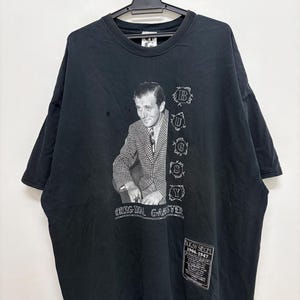 Può includere: T-shirt nero vintage con un'immagine in bianco e nero di un uomo in giacca e cravatta. La maglietta ha la scritta "Bugsy Siegel" e "Original Gangster" stampata sopra. La maglietta è appesa a una gruccia nera.