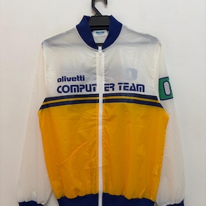 May include: A vintage, zip-up jacket with a white, yellow, and blue color scheme. The jacket features the text "olivetti COMPUTER TEAM" across the chest. The jacket has a blue collar, cuffs, and hem. The jacket is hanging on a black hanger.