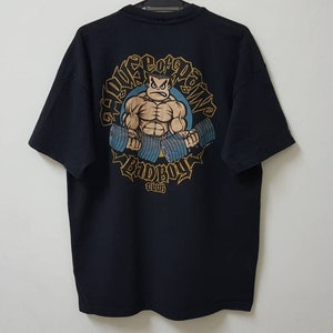 Puede incluir: Camiseta negra con un levantador de pesas de dibujos animados y el texto "HOUSE OF PAIN BADBOY CLUB" en oro y azul. El diseño está en la espalda. La camiseta está hecha de un material suave.