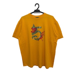May include: Mustard yellow t-shirt with a graphic design. The design features a stylized number 5 with a camouflage pattern and a red and green bird-like figure. The text "Triple Five Soul" is above the number.