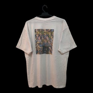 May include: White t-shirt with a colorful graphic on the back featuring a crowd of people. The shirt is hanging on a black hanger against a black background. The graphic appears to be a collage of faces.