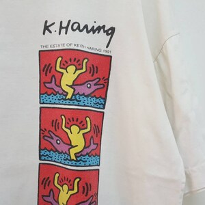K&ouml;nnte beinhalten: Wei&szlig;es T-Shirt mit einem Grafikdruck eines Cartoon-Charakters, der auf einem Delfin reitet. Der Druck ist in Rot, Gelb, Blau und Lila. Der Text "K. Haring THE ESTATE OF KEITH HARING 1991" ist &uuml;ber dem Grafikdruck gedruckt.