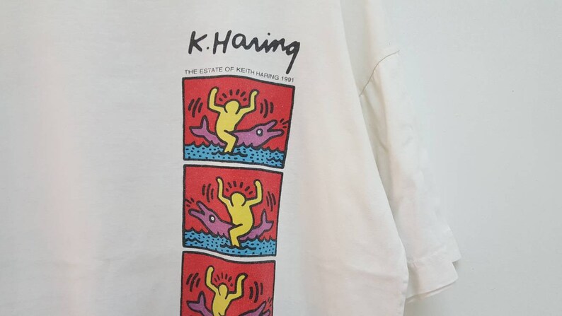 K&ouml;nnte beinhalten: Wei&szlig;es T-Shirt mit einem Grafikdruck eines Cartoon-Charakters, der auf einem Delfin reitet. Der Druck ist in Rot, Gelb, Blau und Lila. Der Text "K. Haring THE ESTATE OF KEITH HARING 1991" ist &uuml;ber dem Grafikdruck gedruckt.